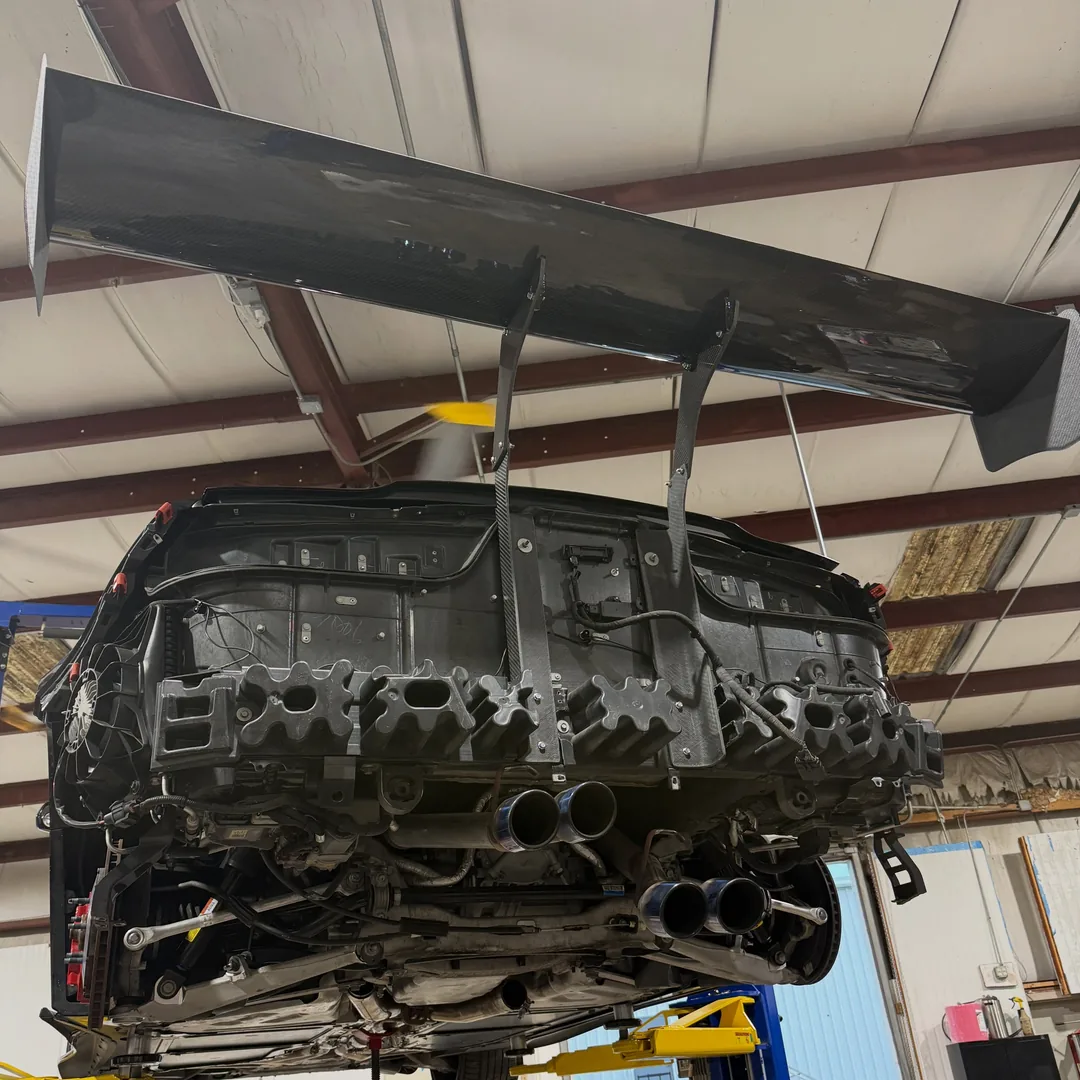 A carbon fibre swan neck wing mounted on a car with the rear bumper removed, up on a lift