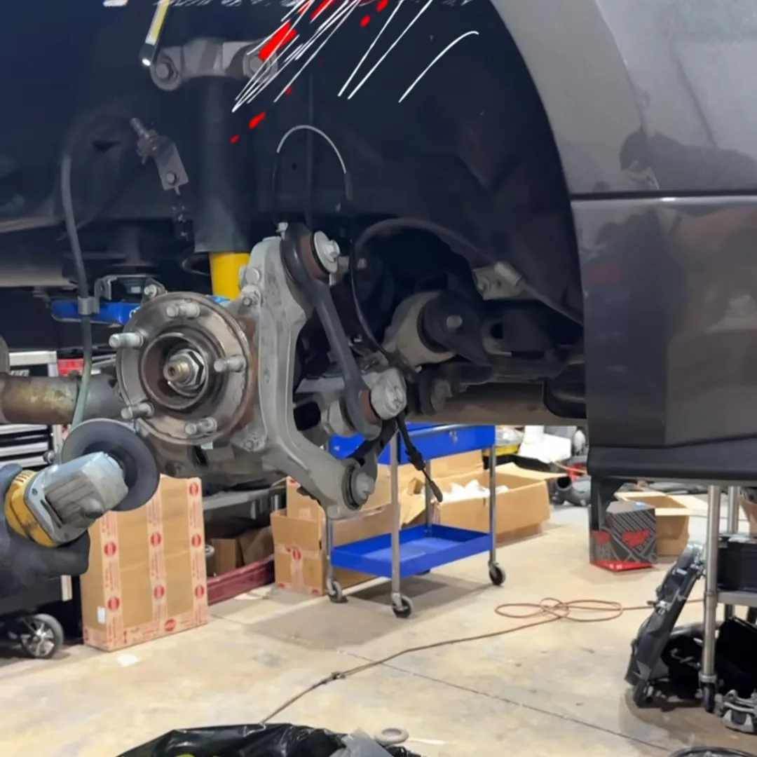A car in the shop with one wheel removed, showing the bare hub, knuckle and suspension, with parts boxes on the floor beside it.