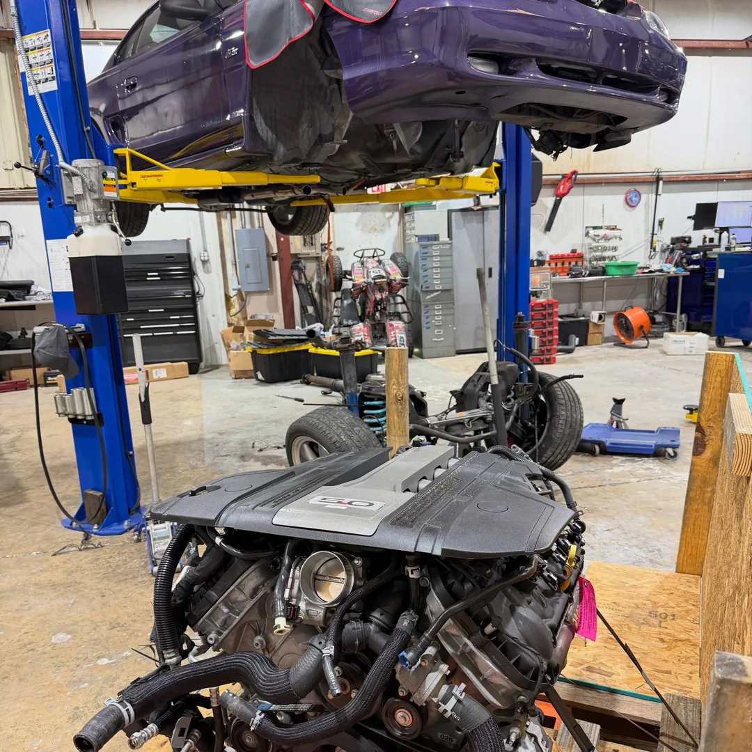 A purple Mustang raised on a blue two post lift with its engine out, and a Coyote V8 sitting on wooden supports in the foreground of the shop.